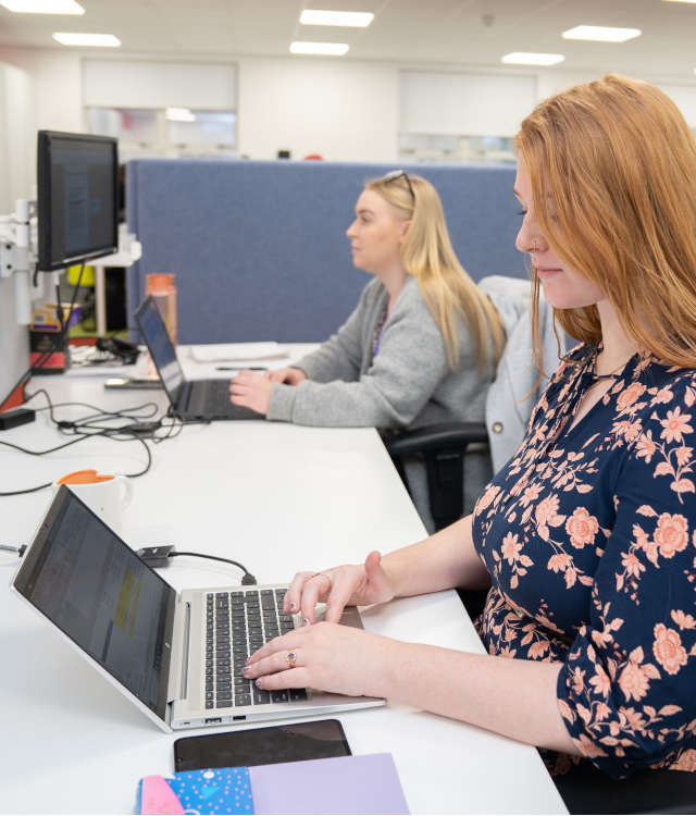 Woman with ginger hair sitting at a laptop, woman with blonde hair on laptop in background