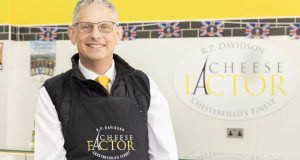 Simon from Cheese Factor in apron in his shop with the company logo on his tiled wall