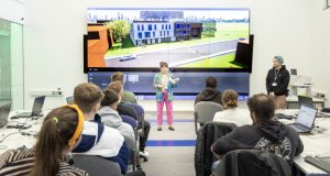 Female lecturer teaching full class, large interactive screen behind lecturer