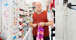 Male in red wool jumper (owner of The Wool Cabin) holding a pink knitted scarf, balls of wool in background