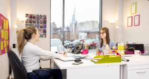 2 female business people at white desks talking at Chesterfield Enterprise Centre, Crooked Spire visible through window