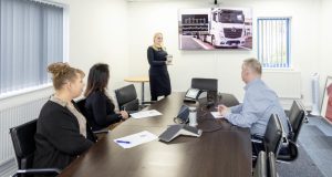 Female standing presenting in front of screen, two females and a male at table listening at Weightron