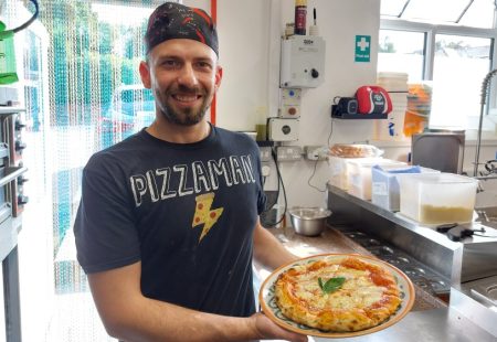 Chef wearing t shirt with pizza man written on the front holding a freshly cooked pizza in kitchen